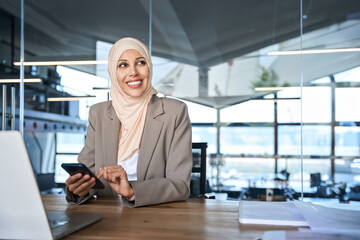 Beautiful portrait of Middle Eastern muslim businesswoman manager ceo using cell phone mobile app. Smiling young indian woman in hijab holding smartphone sitting in office working online. Copy space