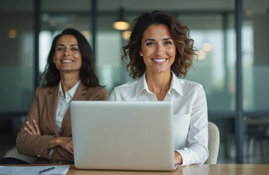 Two smiling businesswomen in modern office. One woman using laptop another folded arms. Middle-aged, hispanic ceo working online, analyzing data, planning business strategy. Successful