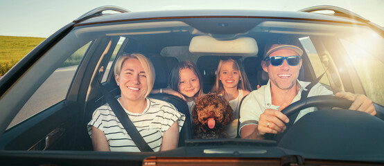 Happy family enjoys road trip together, with fluffy brown Maltipoo dog in front seat and two young...