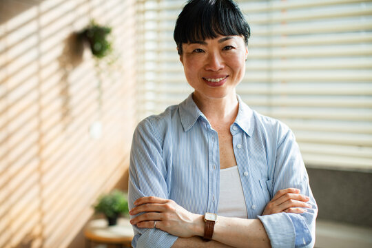 Confident Asian woman smiling with arms crossed while standing in bright modern office