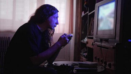 An intensely focused and absorbed mood is captured as a man, illuminated by the blue glow of a screen, plays a video game with a controller in a dimly lit room