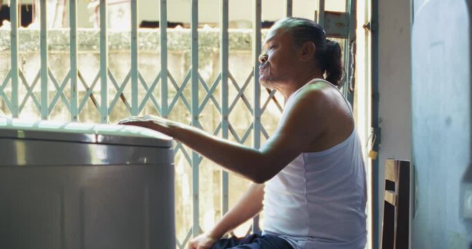 Asian man which is senior, sits beside a washing machine behind his house. Surrounded by concrete and daily items, he does his laundry in silence, his face lost in memory and quiet thought.