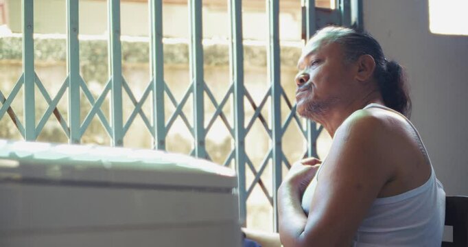 Asian man which is senior, sits beside a washing machine behind his house. Surrounded by concrete and daily items, he does his laundry in silence, his face lost in memory and quiet thought.