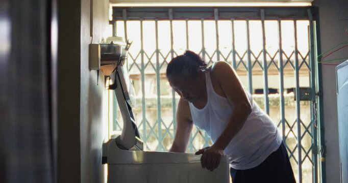 Asian man which is senior, standing beside a washing machine behind his house. Surrounded by concrete and daily items, he does his laundry in silence, his face lost in memory and quiet thought.