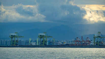 Obraz premium Large port cranes against a cloudy sky with mountains and a city skyline in the background, over calm water