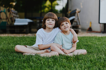 Fototapeta premium Two cheerful boys sitting together on green grass, enjoying a sunny day, showcasing friendship and childhood innocence in a playful atmosphere.