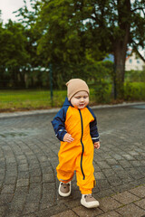 Smiling little boy in rubber boots and blue waterproof overalls having fun playing in a puddle on a rainy autumn day in a park or yard