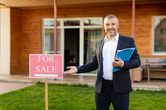 Confident real estate agent with clipboard showing for sale sign, posing in front of modern cottage outdoors