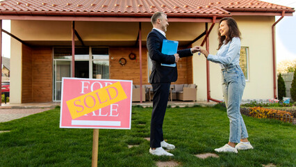 Young woman buying new house, taking keys from male real estate agent, standing outdoors in front of house with sold signboard on foreground