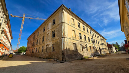 Large historic building with peeling yellow paint and boarded windows stands on a dirt street, with a tall yellow crane and blue sky above, showing signs of restoration work.