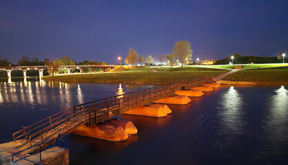 Fototapeta premium Floating pontoon footbridge with orange floats crosses a calm river, glowing under streetlights, with grassy banks and a historic arched bridge lit in the background on a clear night.