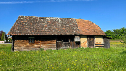 Old wooden barn with a patchwork tiled roof stands in a wildflower-dotted green meadow, showing...
