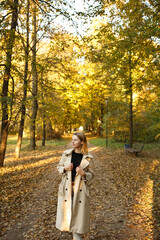 Fototapeta premium Stylish young woman in beige trench coat standing on a forest path during golden autumn sunset. Calm atmosphere, fashion look, casual outdoor lifestyle in nature.