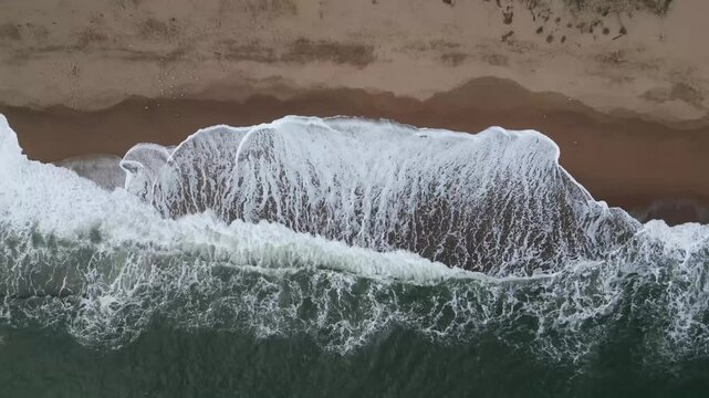 Vista a&eacute;rea en picado desde dron que se eleva sobre playa de Catalu&ntilde;a con olas rompiendo y espuma en la orilla.