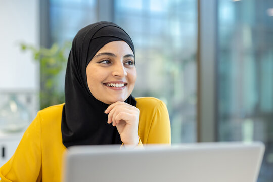 A smiling woman in a hijab is looking off-camera with her hand on her chin. She wears a yellow blouse, and a laptop is in front of her.