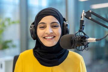A young, smiling woman wearing a hijab and headphones speaks into a microphone, likely recording a...