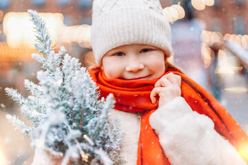 Young child smiles while holding a small snow-covered tree in a festive winter market filled with twinkling lights during a snowy day