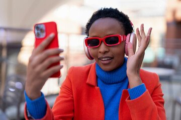 Woman wearing red sunglasses and headphones enjoys music while taking a selfie in a vibrant urban setting during bright daylight hours
