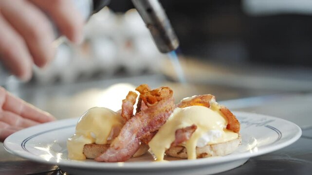 A plate of eggs benedict with bacon and a person preparing it. The plate is on a table and the person is wearing gloves