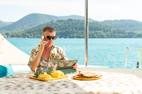 Caucasian businessman working on digital tablet and speaking on mobile phone for online corporate business on luxury catamaran boat yacht sailing in ocean. Workation on summer beach holiday vacation.