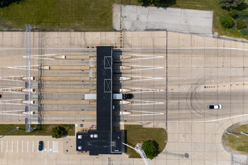 Top-down aerial view of a highway toll plaza with multiple lanes, a central toll booth building, and vehicles including a semi-truck passing through under bright daylight in a suburban area.