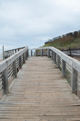 Wooden bridge guiding visitors toward the beach under a gray, cloudy sky.

