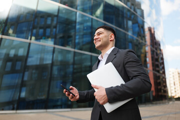 Young Professional Smiles Confidently While Walking Outside a Modern Office Building With Laptop...