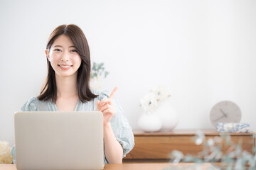 自宅の部屋でパソコンを使う若いかわいいカジュアルな服の女性カメラ目線　ポイントやオススメの指差しポーズ