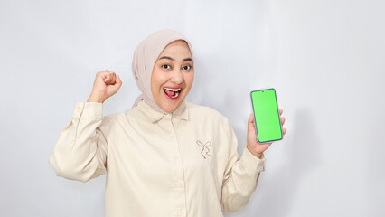 Beautiful Asian Muslim woman wearing hijab with happy expression while holding her smartphone and clenching her fist facing up. on white background.