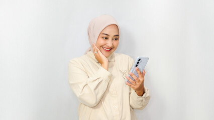 Beautiful Asian Muslim woman wearing hijab smiling while looking at her smartphone screen on white background.
