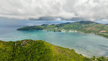 Dense tropical forest-covered hills with a calm bay and distant village in the background. Praslin, Seychelles.