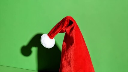 Red Santa hat against a solid green background, casting a stark shadow, symbolizing the holiday season and festive spirit. - Powered by Adobe
