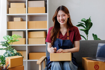 Young asian woman checking supplies and writing address on box with laptop in her warehouse. Owner,...