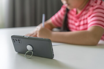 closeup smartphone with Asian child or kid girl student writing or drawing on paper by left hand to study online learning by mobile phone in classroom homeschool or people learn from home and studying
