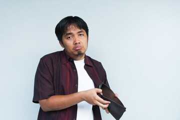 Asian Indonesian man showing his empty wallet with sad and worried expression. Isolated in gray background.