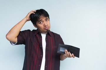 Asian Indonesian man showing his empty wallet with sad and worried expression. Isolated in gray background.