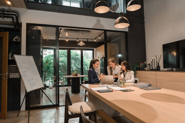 A group of three young busineewoman are sitting at a desk with documents, tablet, laptop, and other equipments. They are having a discussion and planning session to improve the company.