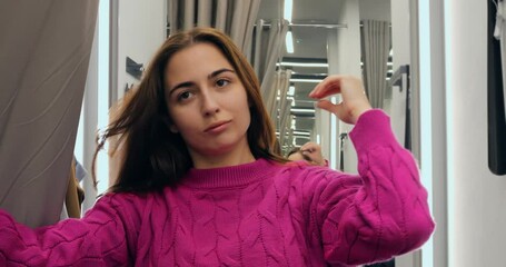 Stylish female checking reflection while trying on vibrant fuchsia sweater, adjusting hair and posing confidently inside clothing store dressing room. Concept of buying women clothes - Powered by Adobe