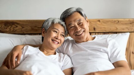Elderly mature Asian couple embracing, smiling, relaxing in bed together at home, enjoying retirement, love, bonding, and healthy aging - Powered by Adobe