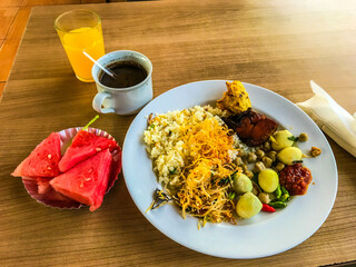 Plate of Indonesian breakfast with rice, vegetables, sambal, grilled chicken, coffee, orange juice, and fresh watermelon slices on a wooden table.

