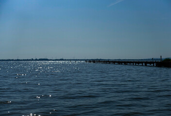 A jetty stretches across a calm, sunny lake, surrounded by clear skies. The Dümmer, also referred...