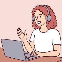 Young woman in headset works remotely on laptop and waves hand while talking during online meeting or video call in casual home setting