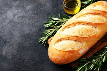 A loaf of crusty bread on a wooden board with fresh rosemary sprigs and a small glass jar of olive oil on a dark textured surface.