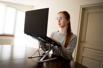 Portrait of young woman with eyeglasses working on laptop at adjustable laptop stand for desk for good posture at home office, female using computer with ergonomic workspace