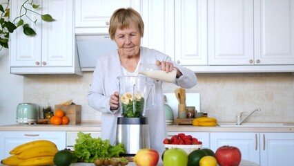 Elderly woman adding plant based milk to a blender filled with spinach, banana, and other ingredients, preparing a nutritious smoothie in her modern white kitchen surrounded by fresh produce