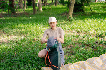 Woman unpacking a camping bag on a sunny day in a green park surrounded by trees while sitting on a picnic blanket