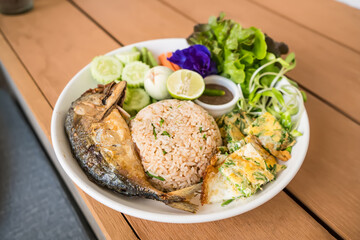 Delicious Thai rice dish with fried fish, omelette, fresh vegetables, and dipping sauce served on a white plate on a wooden table