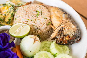 Delicious Thai rice dish with fried fish, omelette, fresh vegetables, and dipping sauce served on a white plate on a wooden table