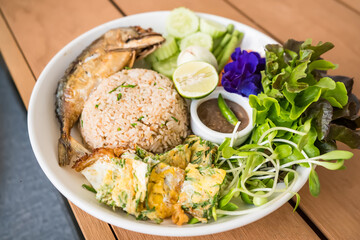 Delicious Thai rice dish with fried fish, omelette, fresh vegetables, and dipping sauce served on a white plate on a wooden table
