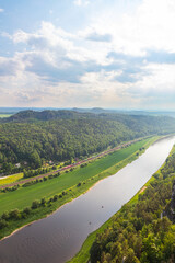 Scenic panoramic view from the Bastei cliffs over the Elbe River valley with sandstone rocks, lush forests, rural village rooftops, distant mountain ranges under a vibrant summer sky. Saxony, Germany